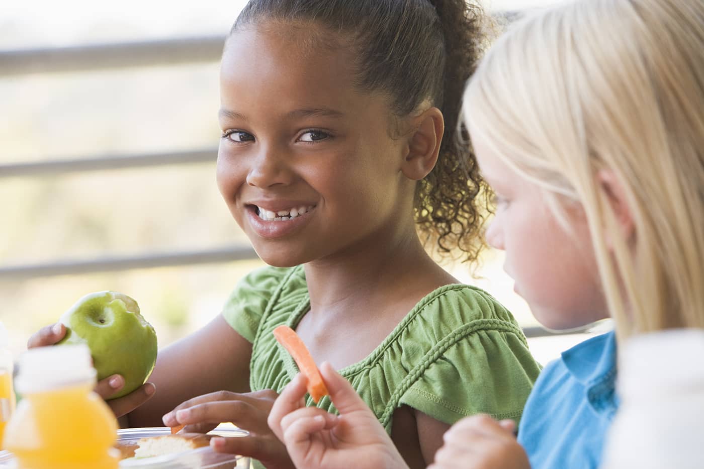 Two young girls, one smiling at the camera, sit next to each other while eating nutritious snacks.