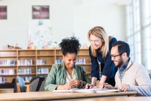 3 adults sit at a table together in a library collaborating on a project