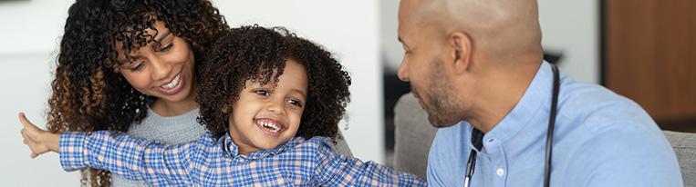 A smiling young child on a mother's lap talks with a health care provider.