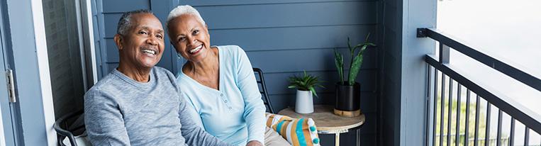 An older couple holds hands and smiles while sitting on their porch.