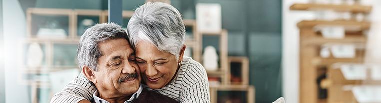 An older man sits on a couch while his wife leans over the back of the couch and hugs him.