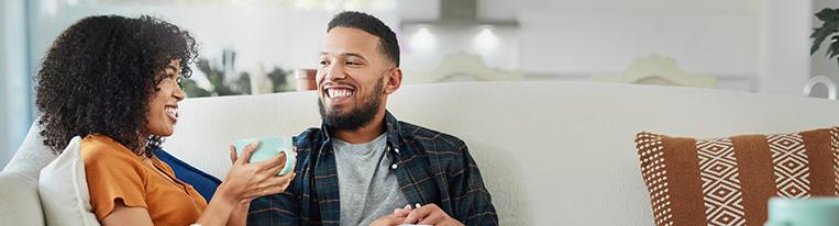 A woman holding a mug sits sideways on a couch with her legs across a man's lap while they look at each other and smile.