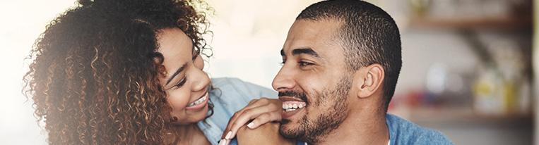 A woman standing next to a man puts her hands on his shoulder while they look at each other and smile.