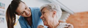 A woman has her hands on an older woman's shoulders as they smile at each other.