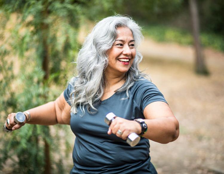 A woman with long gray hair smiles while exercising outside with two dumbbells.