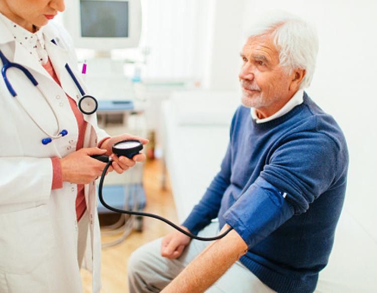 A man with white hair wearing a blue sweater has his blood pressure checked by a doctor wearing a white lab coat.