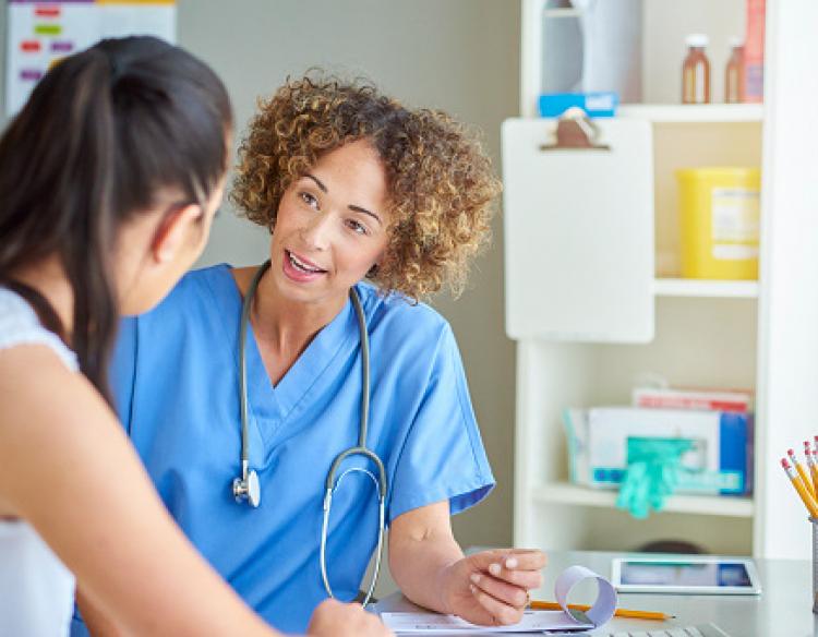 A nurse with dark curly hair wearing blue scrubs talks to a patient.