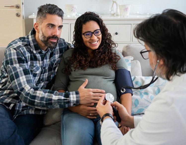 A pregnant woman and partner sit on a couch while a clinician wearing white lab coat checks the pregnant person’s blood pressure.