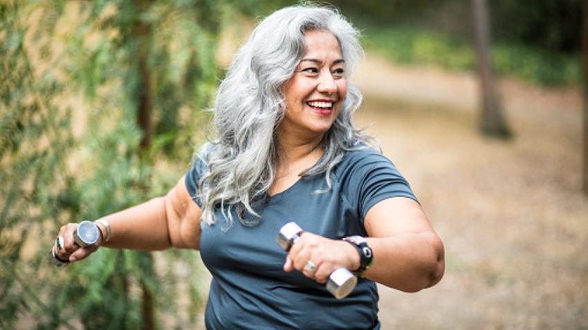 A woman with long gray hair smiles while exercising outside with two dumbbells.