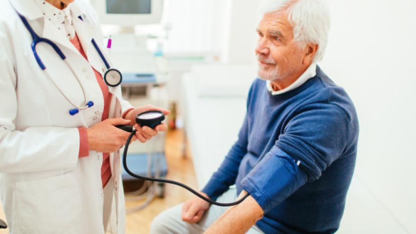 A man with white hair wearing a blue sweater has his blood pressure checked by a doctor wearing a white lab coat.