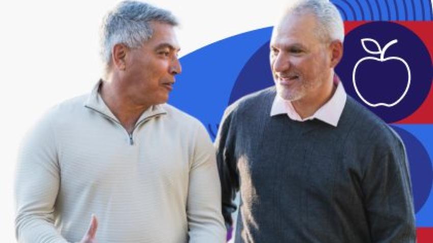 Two older men walk and talk together while one places his hand on the others back.